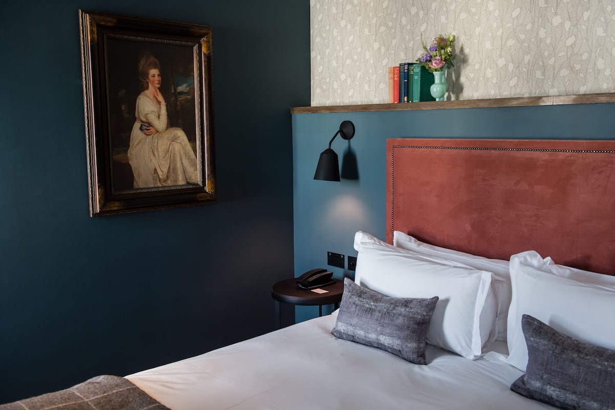 A hotel room with a bed, a framed portrait, a bedside lamp, a small table with a phone, and a shelf with books and flowers.