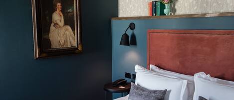 Une chambre d’hôtel avec un lit, un portrait encadré, une lampe de chevet, une petite table avec un téléphone et une étagère avec des livres et des fleurs.