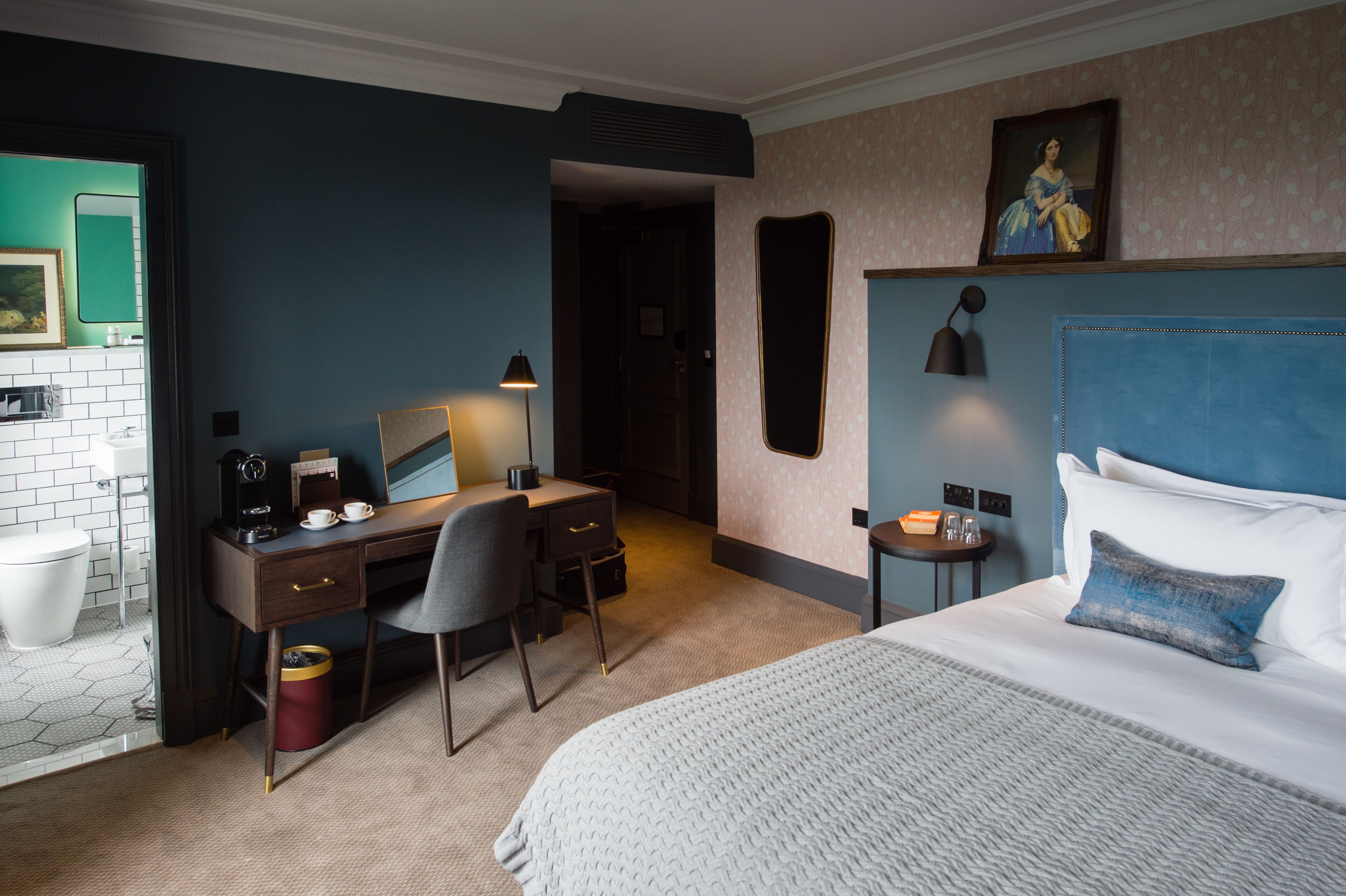 A hotel room with a bed, desk, chair, and a small table with a lamp.