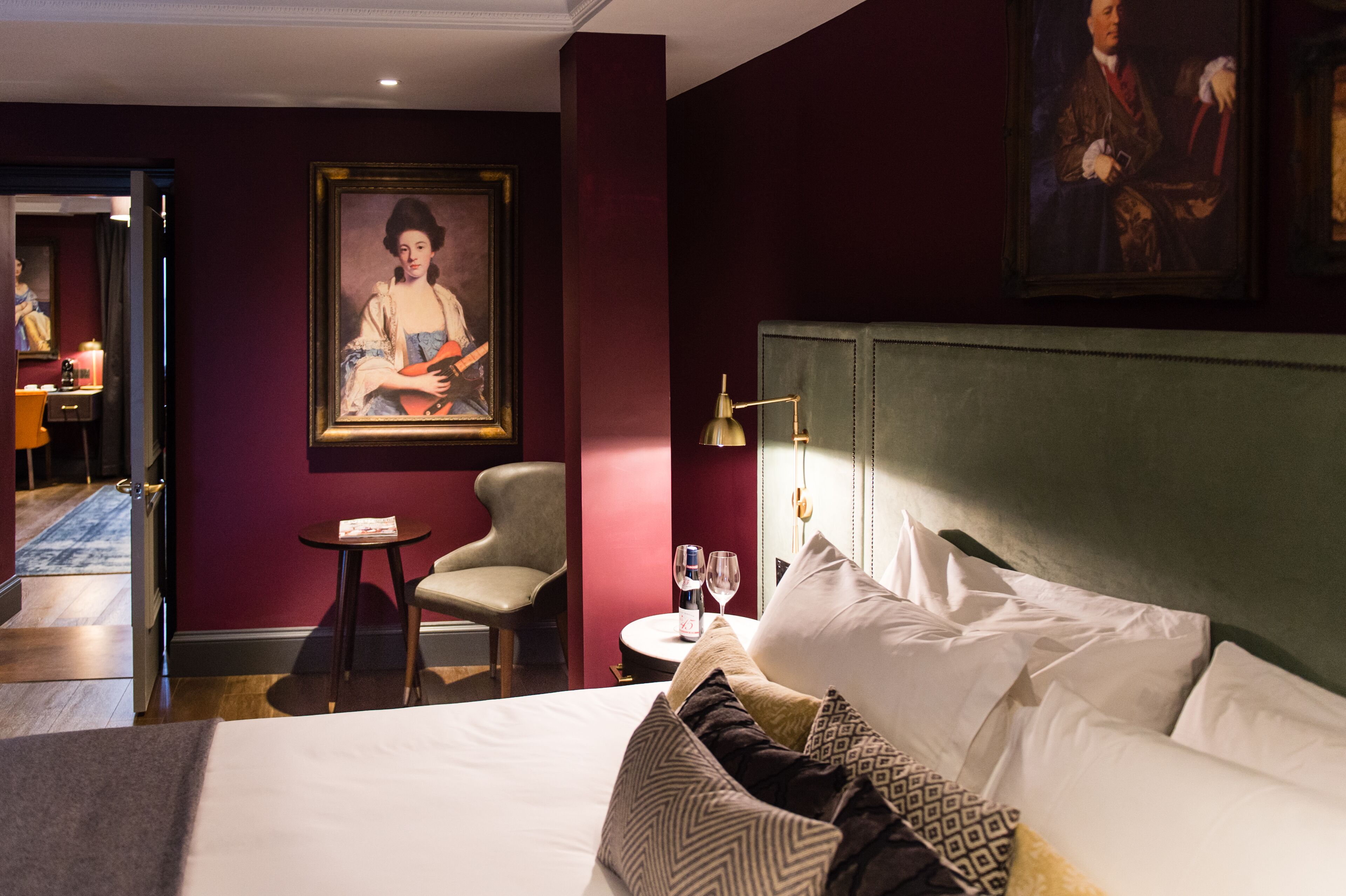A hotel room with a large bed, a chair, a small table with a bottle and glasses, and a painting on the wall.