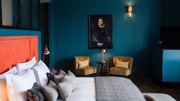 Een hotelkamer met een groot bed, twee fauteuils, een klein tafeltje, een televisie en een ingelijst schilderij aan de muur.