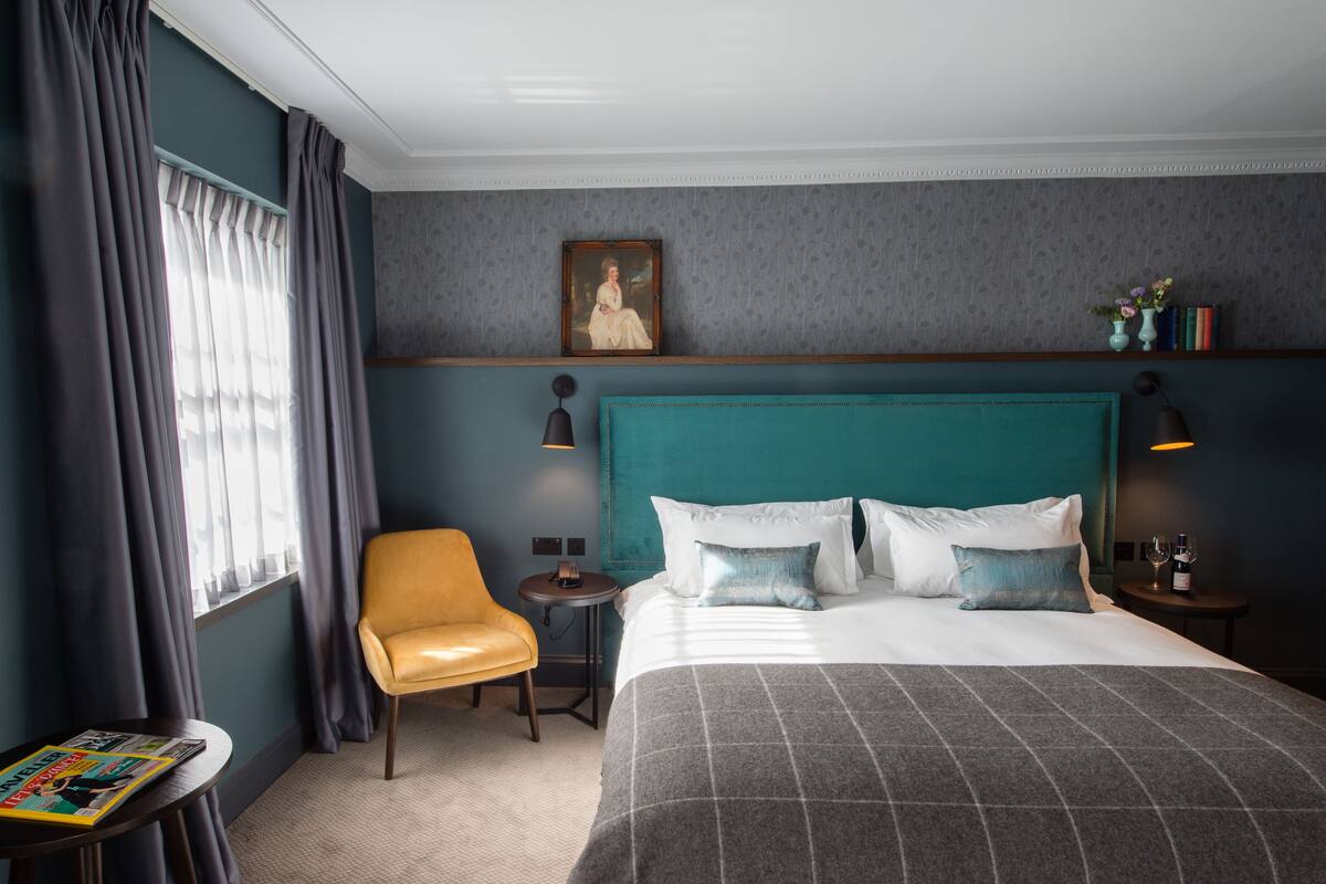 A hotel room with a large bed, a chair, a small table, and a nightstand with a lamp.
