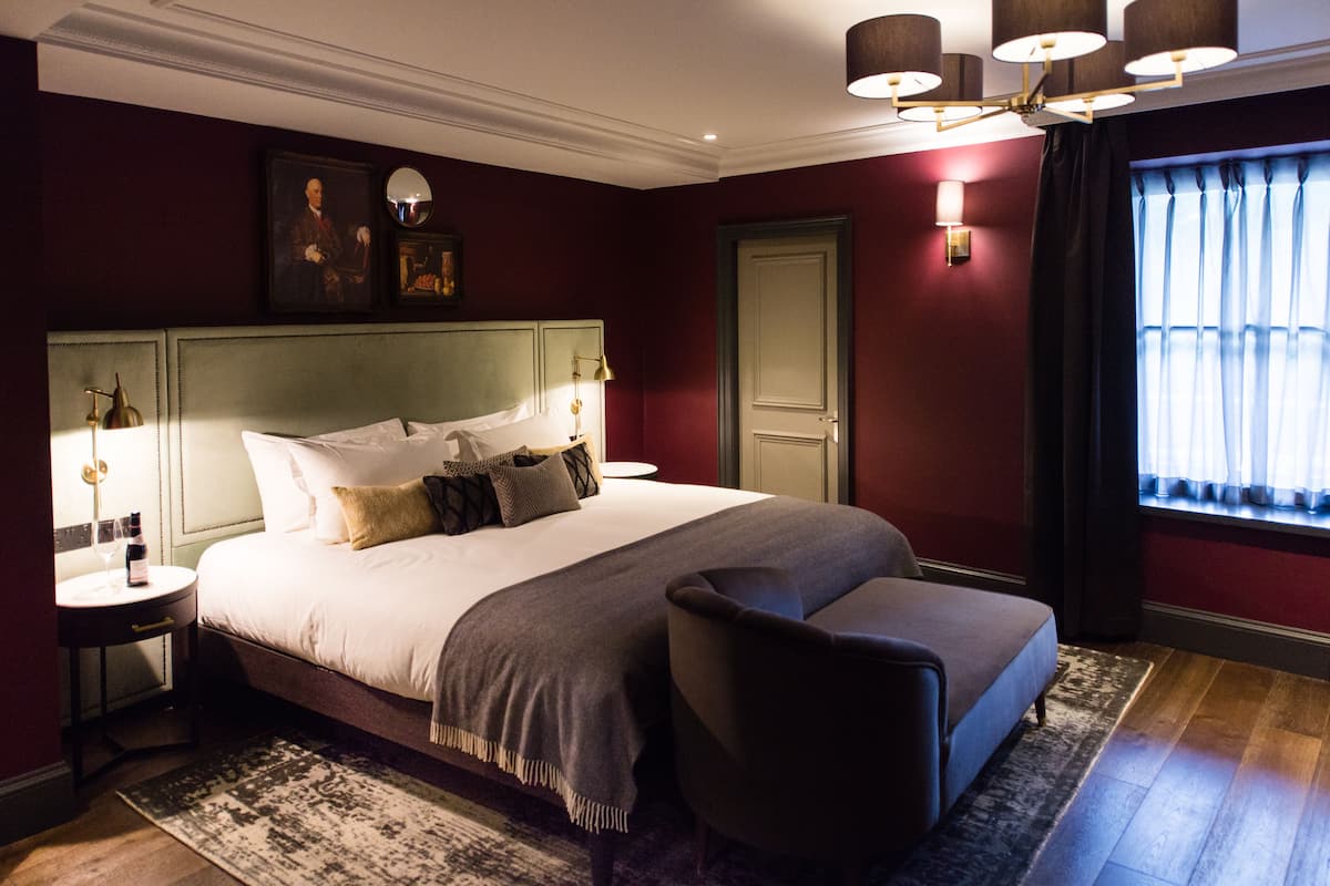 A hotel room with a large bed, bedside tables, a chair, a window with curtains, and a wall-mounted mirror.