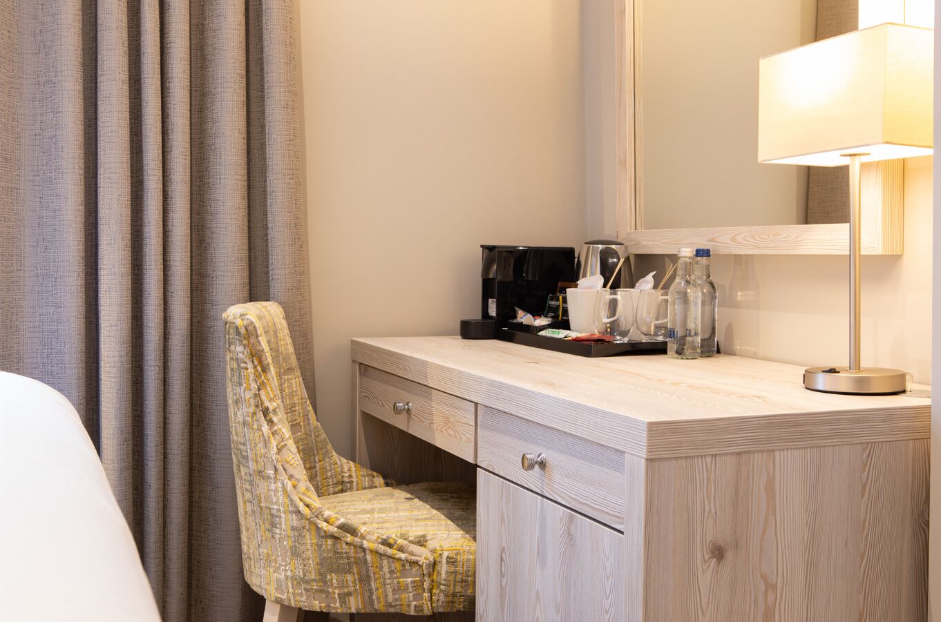 A hotel room with a wooden desk, a chair, a coffee maker, and a lamp.