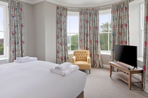 Une chambre d’hôtel avec un grand lit, une chaise, un téléviseur posé sur un meuble en bois et des rideaux à motifs floraux.