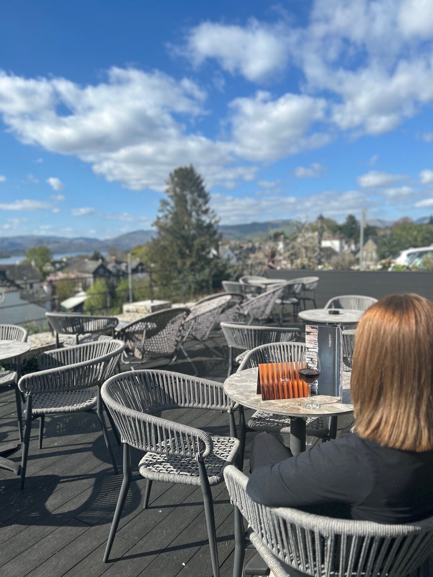 An outdoor seating area with wicker chairs and tables, overlooking a scenic landscape with hills and a body of water.