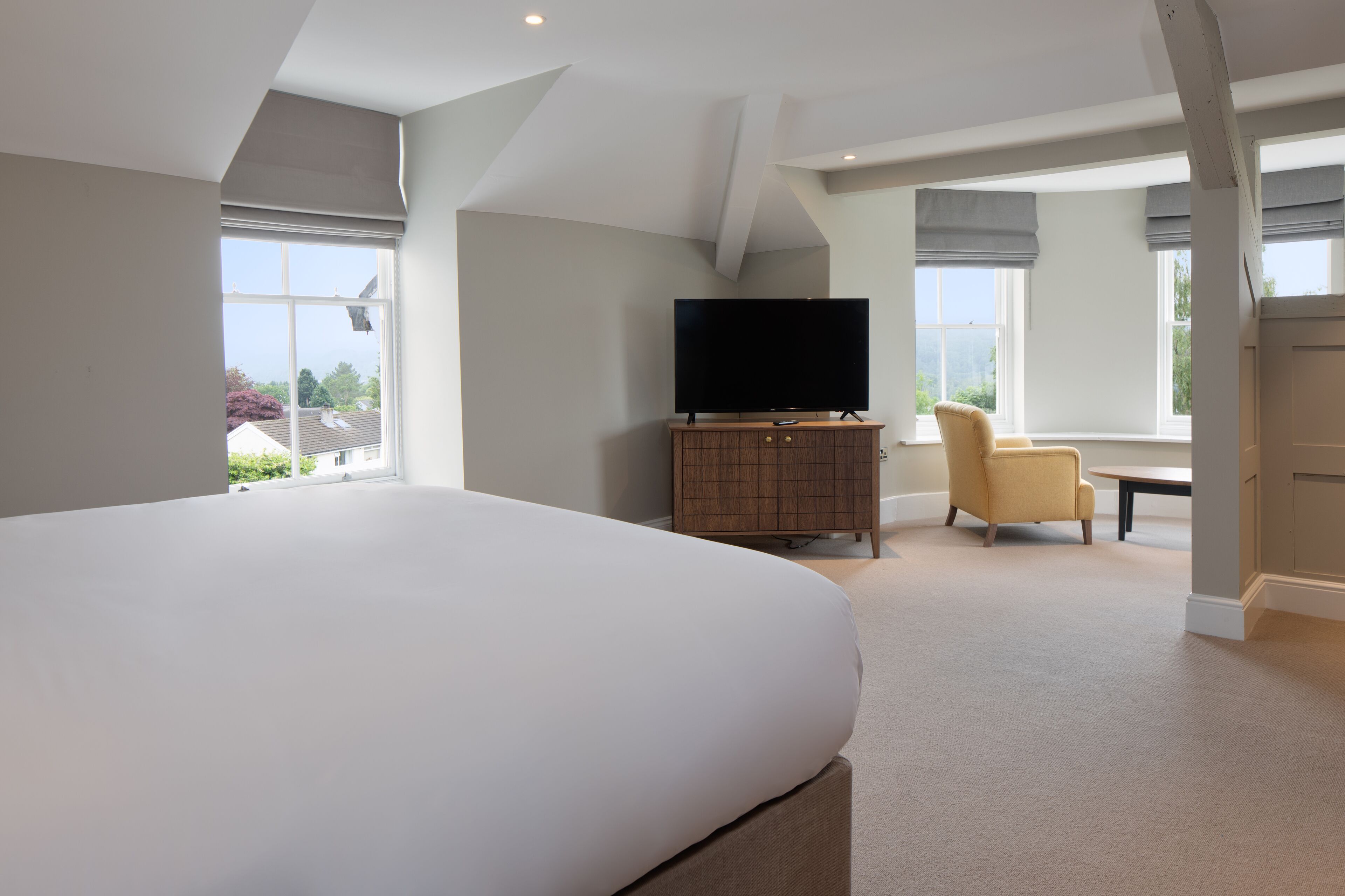 A modern hotel room with a large bed, a flat-screen TV on a wooden cabinet, a comfortable armchair, and large windows offering a view of the outside.