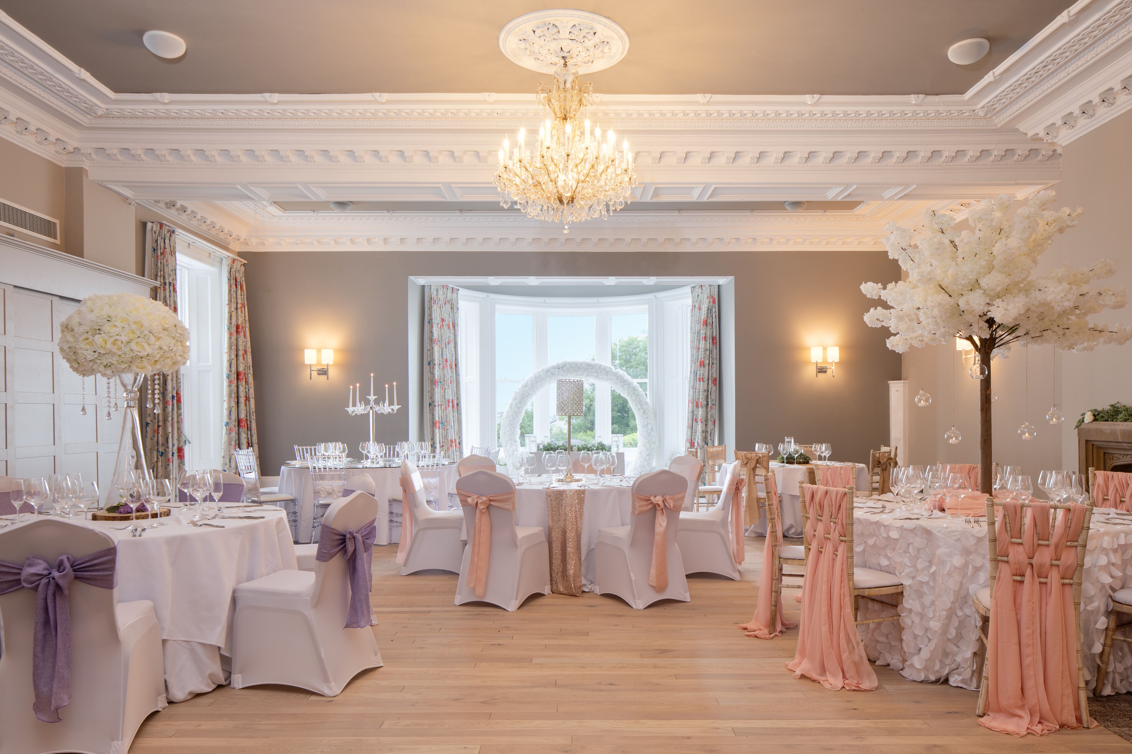 A formal dining room with multiple tables set for a banquet, chandeliers, and large windows.