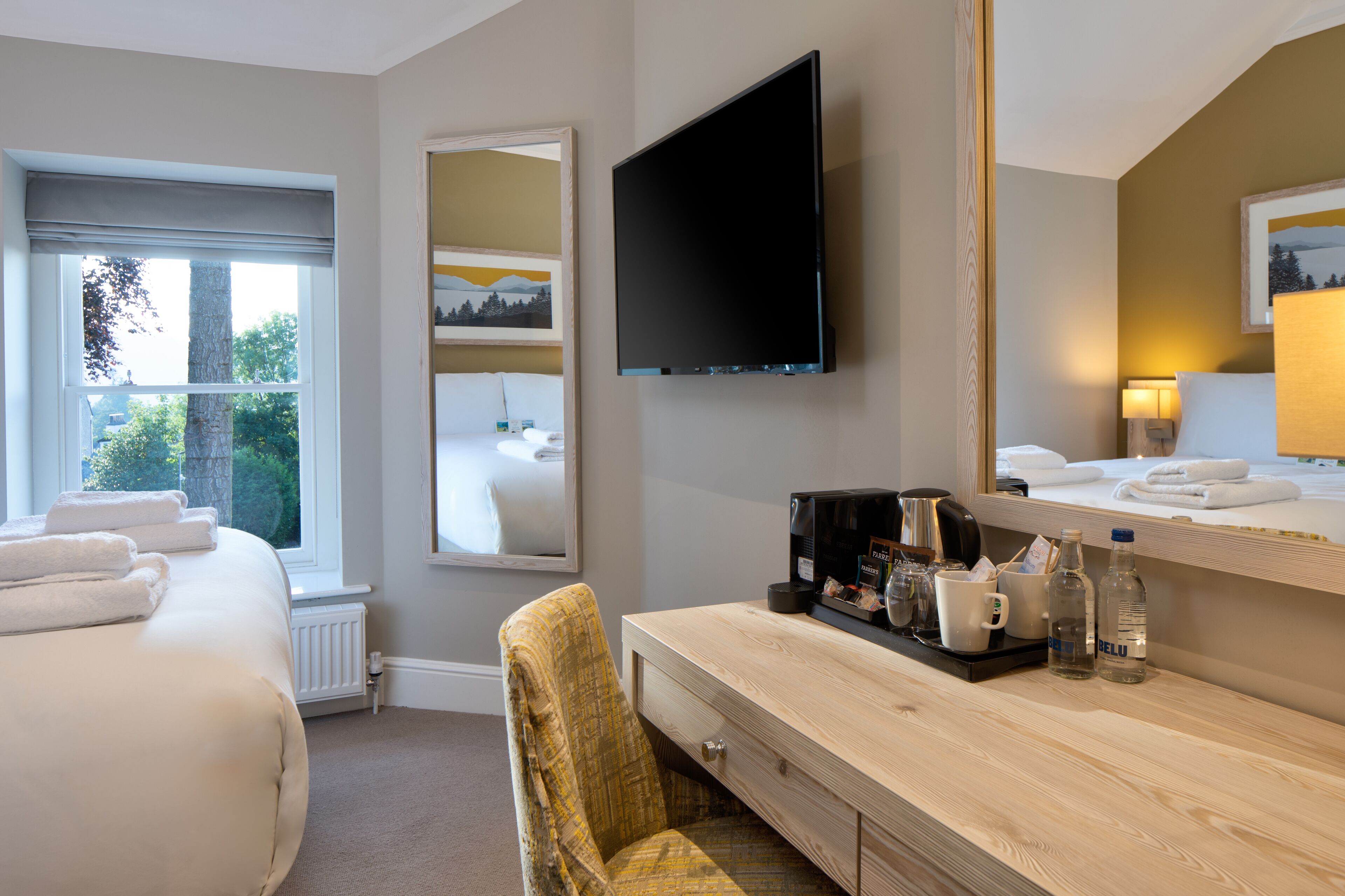 A hotel room with a bed, a desk with a mirror, a television, and a window with towels.