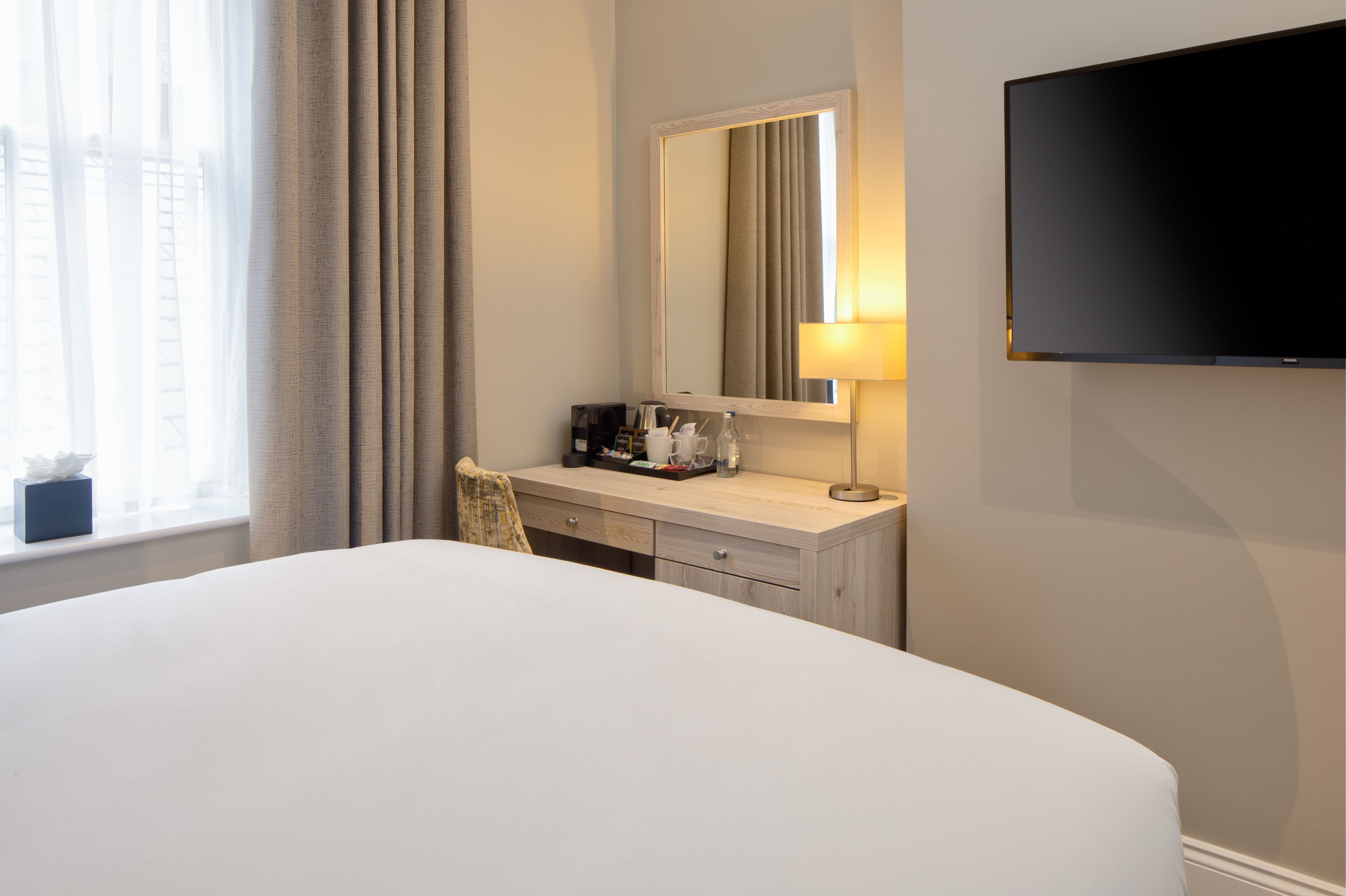 A hotel room with a large bed, a desk with a lamp, a flat-screen TV, and a window with curtains.