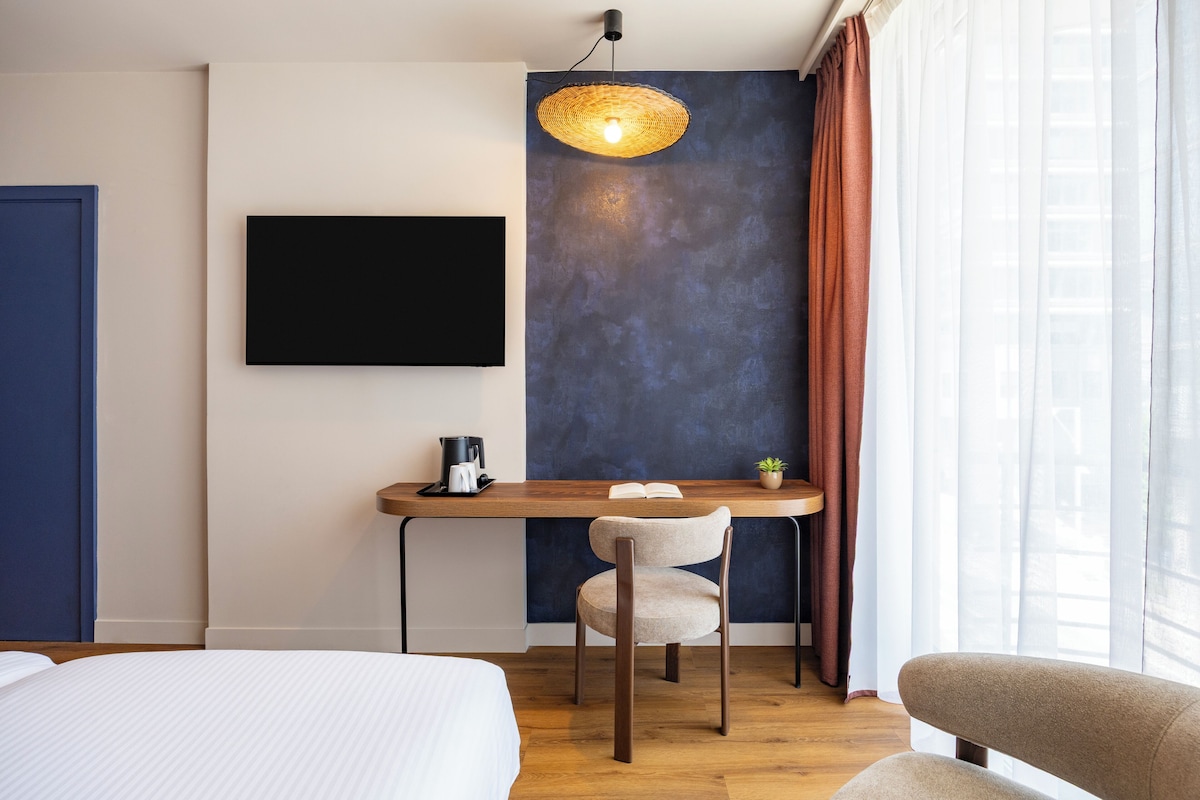 A modern hotel room with a bed, a desk with a chair, a television, and a large window with curtains.