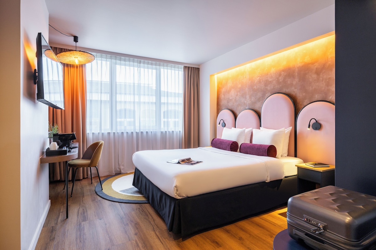 A modern hotel room with a large bed, a desk with a chair, a suitcase, and a wall-mounted lamp.