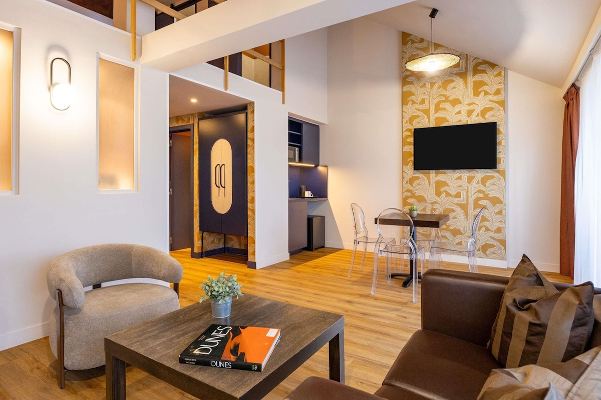 A modern living room with a sofa, a coffee table with a book, a dining area with chairs, a kitchen area, and a TV mounted on the wall.
