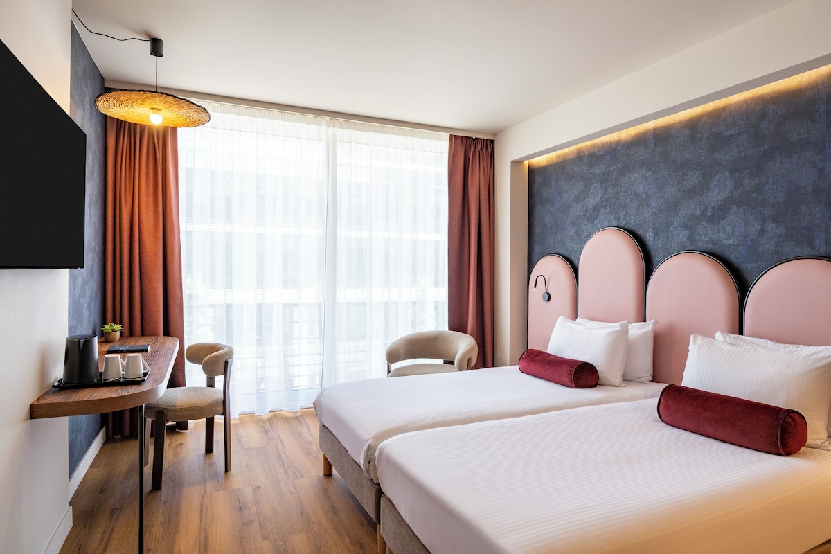 A hotel room with a bed, bedside tables, a desk, a chair, and a large window with curtains.