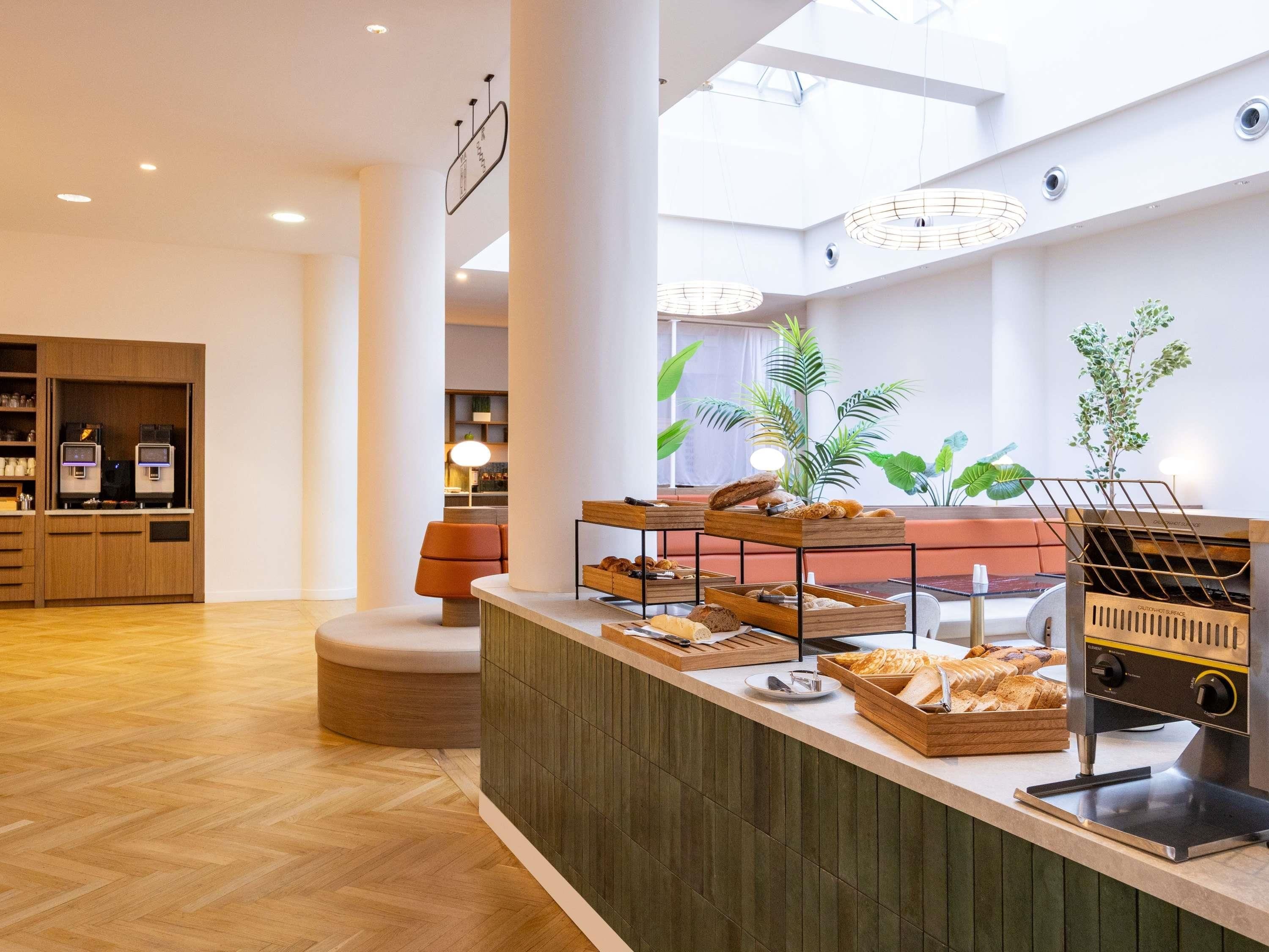 A modern interior with a central counter displaying various baked goods, a seating area with a cushioned bench, and a visible coffee machine.