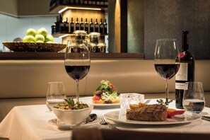 A dining table set with a salad, steak, and two glasses of red wine, with a wine bottle and a basket of green apples in the background. - Estrel Hotel Berlin (Berlin)