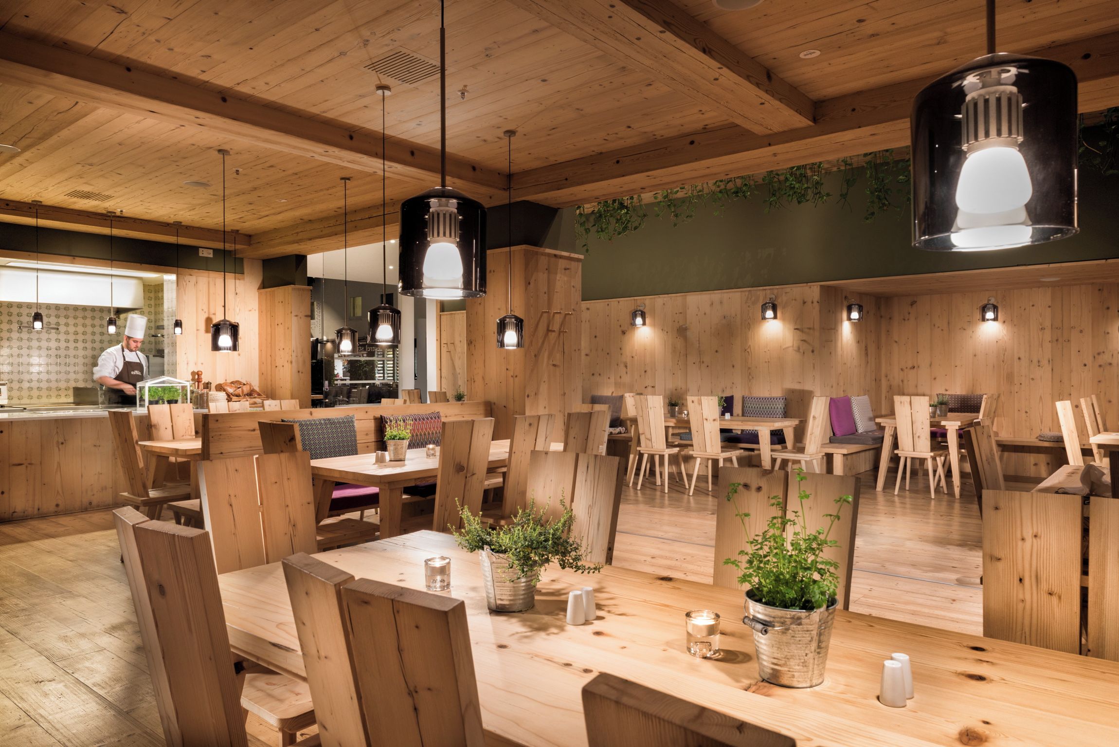 A restaurant interior with wooden furniture, hanging pendant lights, and a chef at the counter.