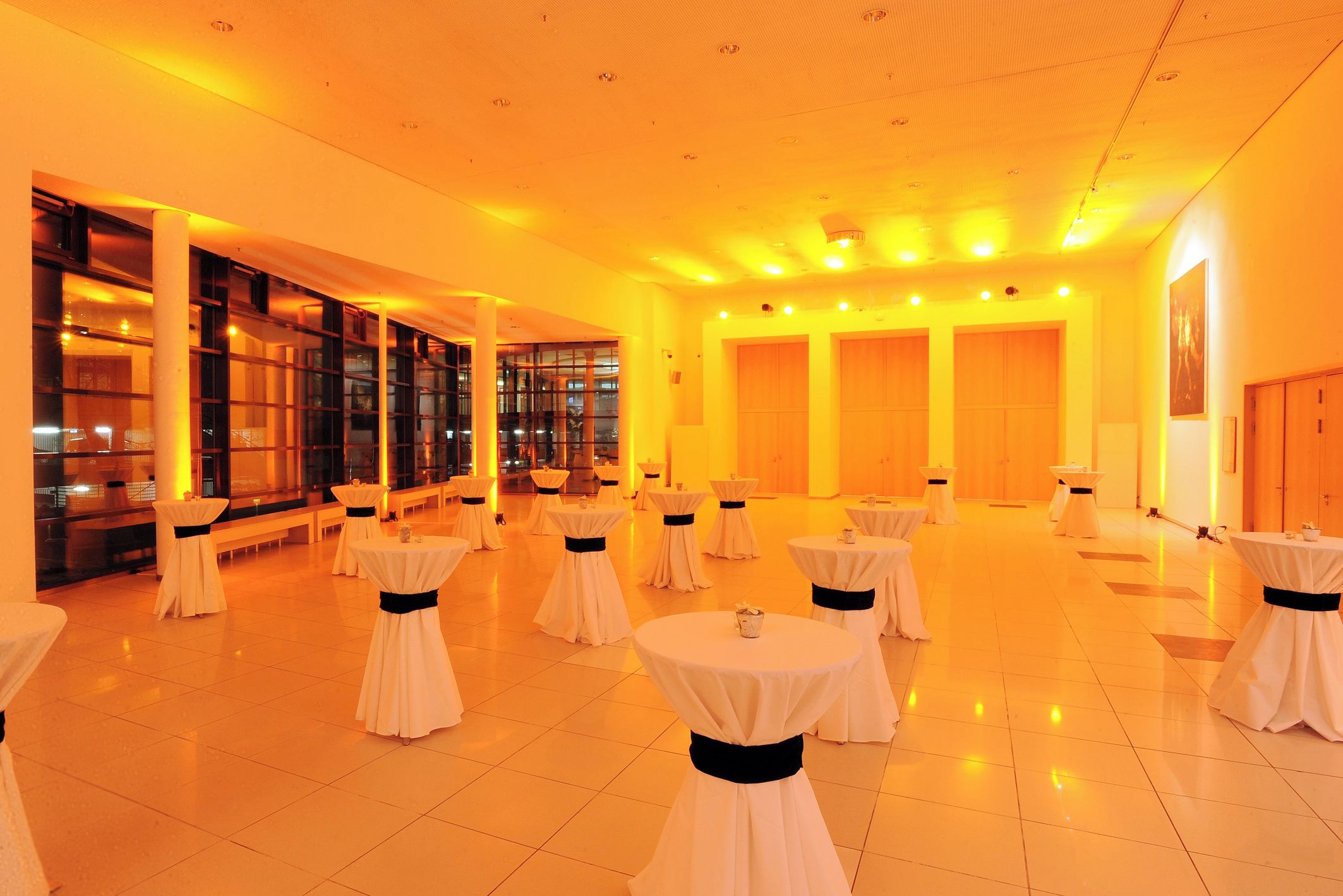 A spacious hall with multiple round tables, each with a white tablecloth and a black sash, arranged in rows.