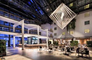 Um átrio amplo, com área de bar central, vários espaços de estar e uma luminária geométrica marcante.