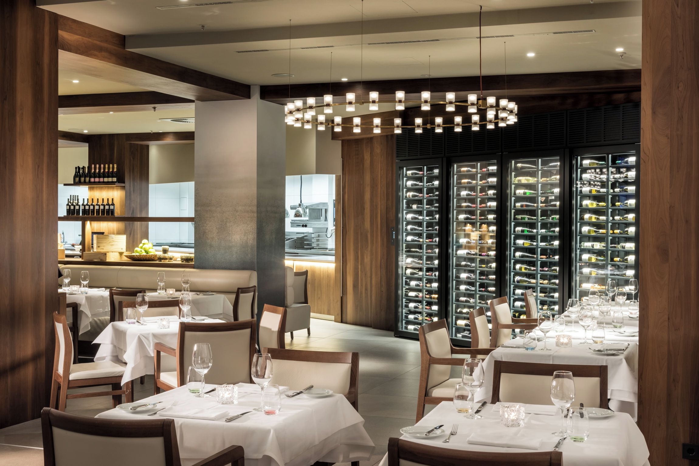 A restaurant interior with multiple dining tables, chairs, and a wine cellar visible through glass doors.