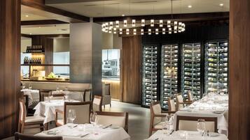 A restaurant interior with multiple dining tables, chairs, and a wine cellar visible through glass doors.