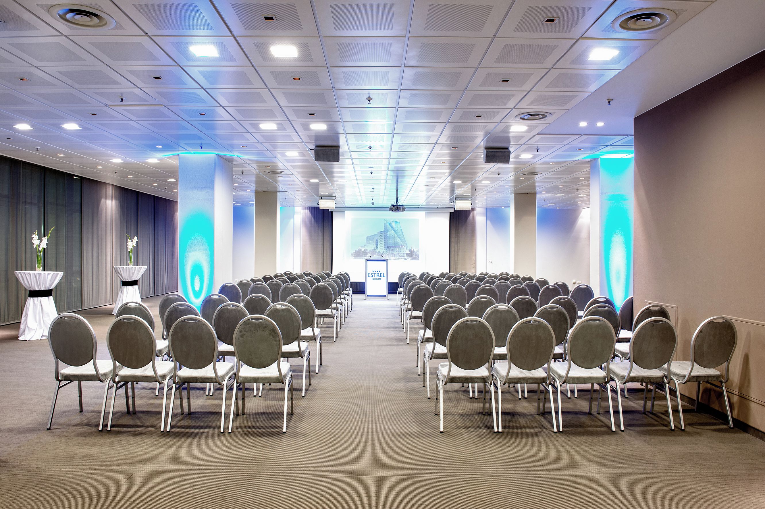A conference room with rows of chairs facing a stage, equipped with a large screen and a podium.