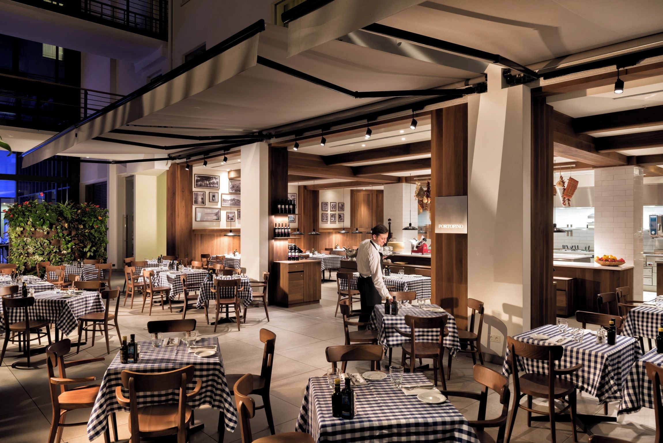 An indoor dining area with checkered tablecloths, wooden chairs, and a bar area with a chef preparing food.