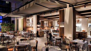 An indoor dining area with checkered tablecloths, wooden chairs, and a bar area with a chef preparing food.
