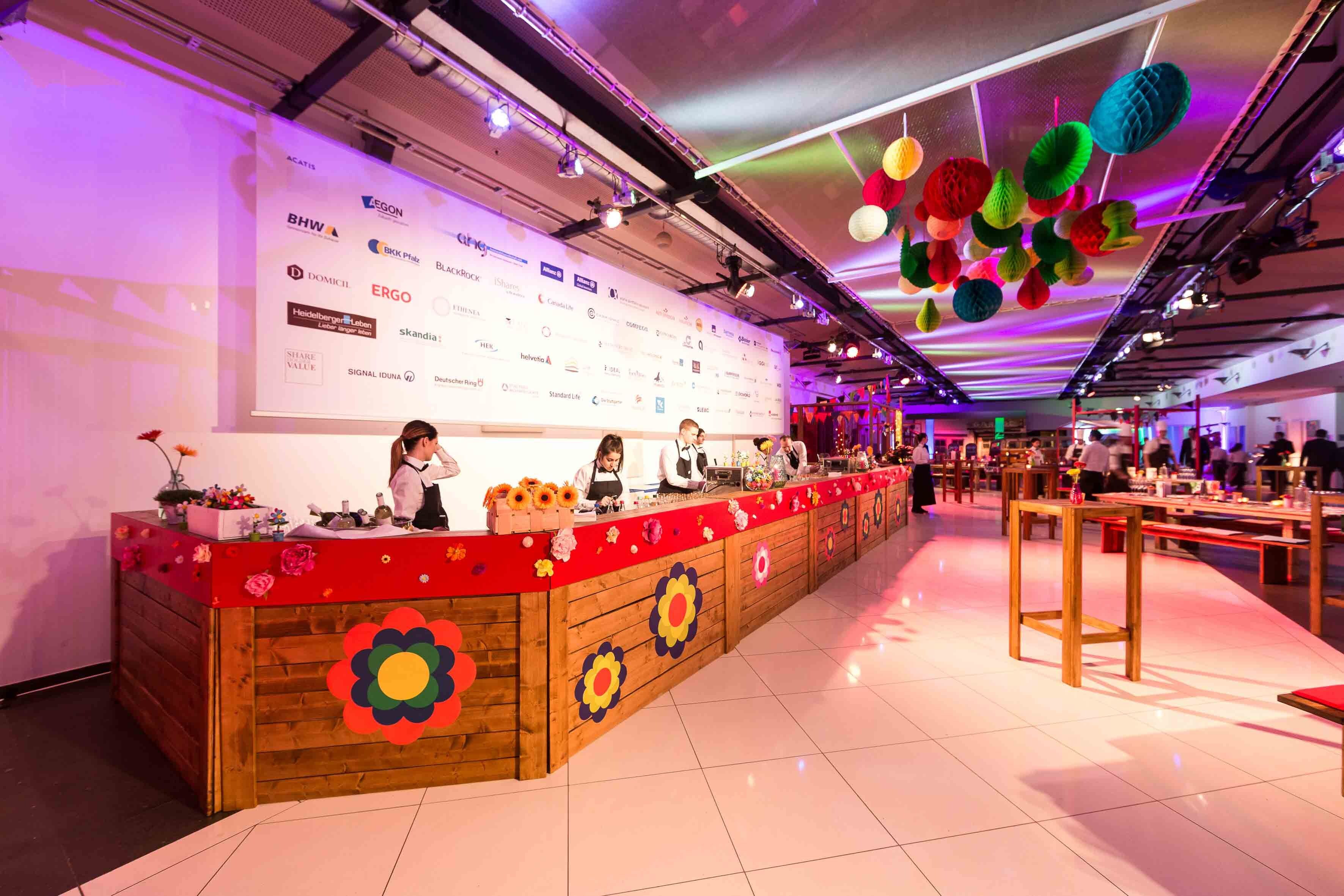 Un espace d’accueil doté d’un comptoir en bois, de décorations florales et d’un mur orné des logos des commanditaires.