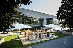 An outdoor seating area with tables and chairs, umbrellas, and a water feature in front of a modern building. - Estrel Hotel Berlin (Berlin)