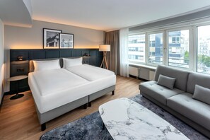 Een moderne hotelkamer met een groot bed, een bank, een klein tafeltje en uitzicht op de stad door grote ramen.