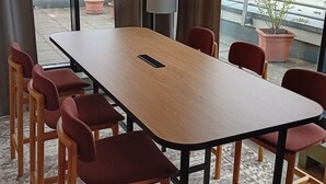 Un salón de conferencias con una larga mesa y seis sillas, ventanas amplias con cortinas, y vistas a edificios y zonas verdes al exterior.