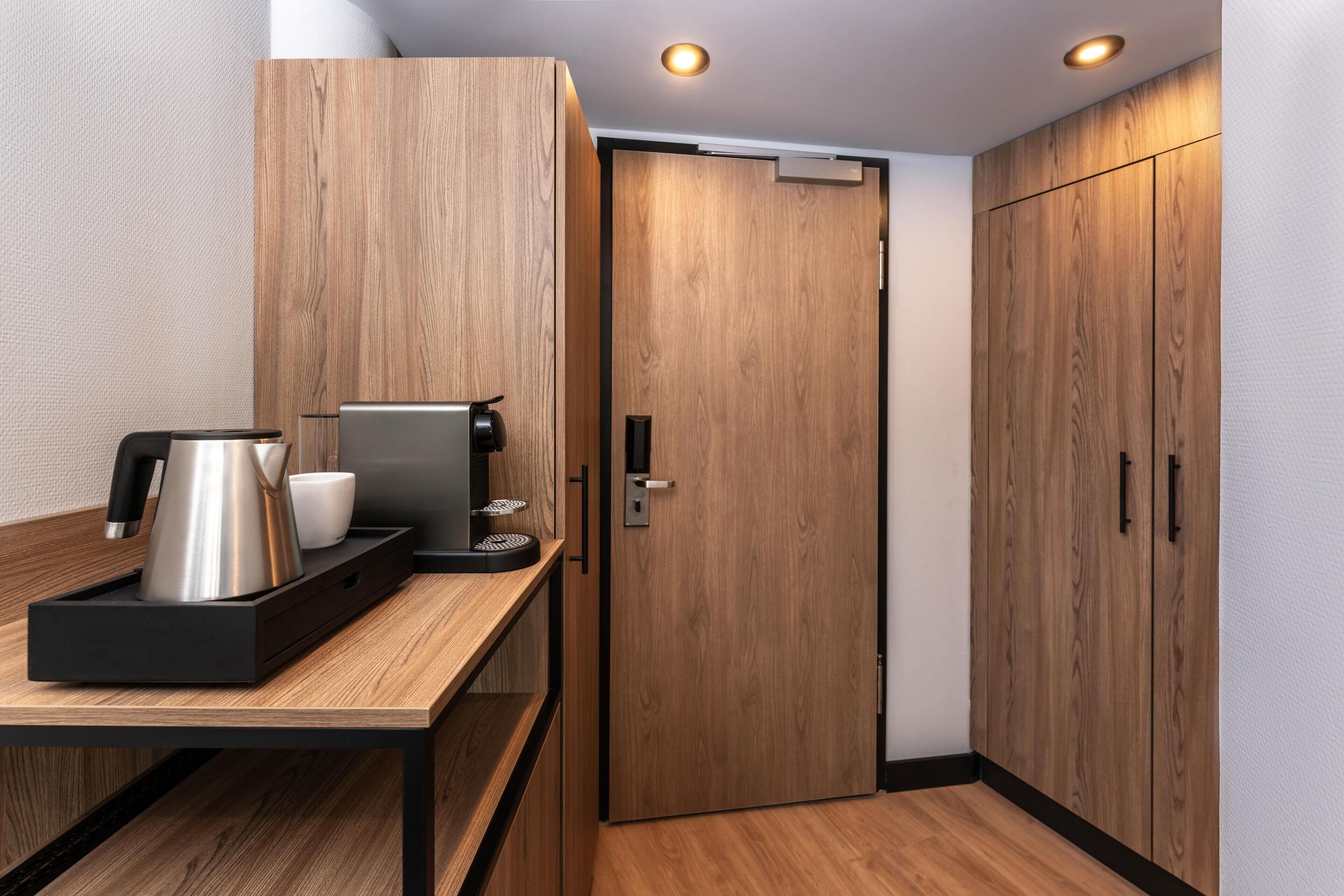 Moderni hotellihuone, jossa on puinen vaatekaappi, kahvinvalmistuspaikka ja ovenkahvallinen ovi.