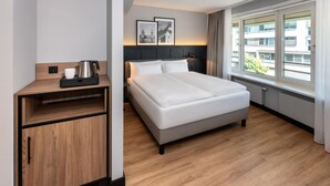 Habitación de hotel con cama, mesita de noche y ventana con vista a edificios.