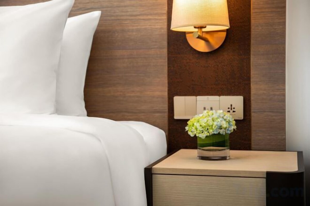 Una cama bien tendida con sábanas blancas, una mesita de noche con un florero y una lámpara de pared.
