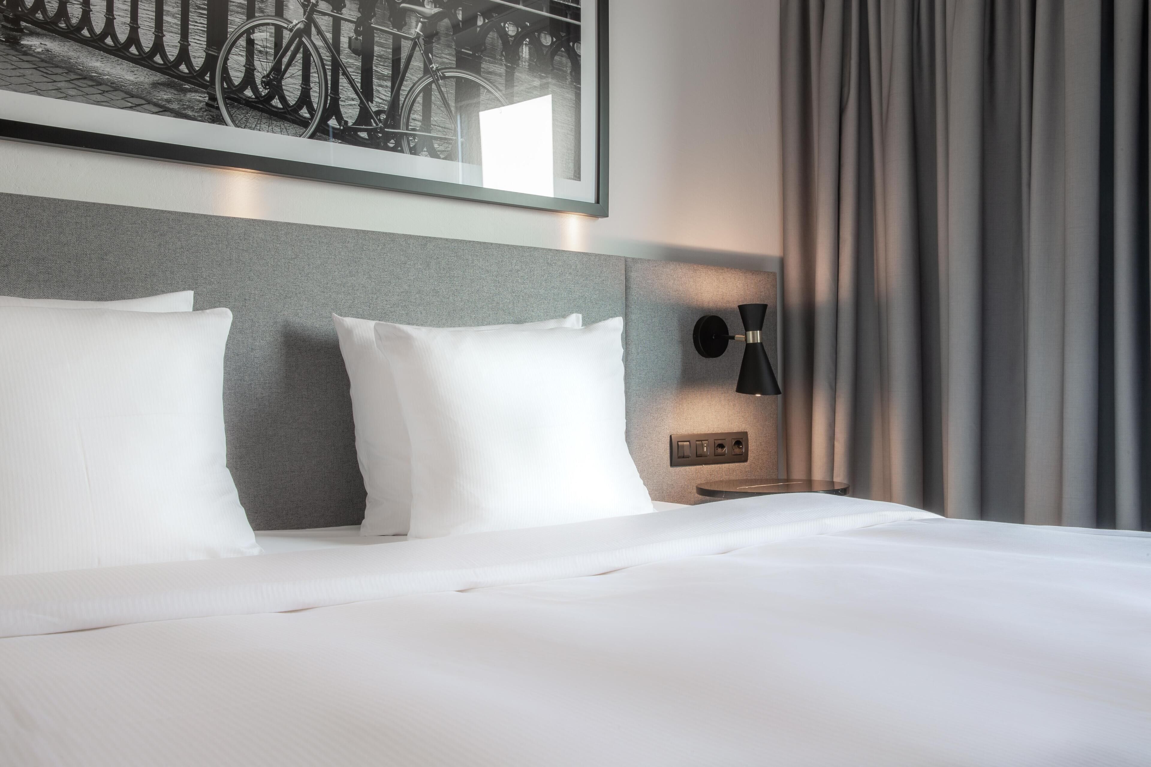 Habitación de hotel con una cama bien hecha, cabecera con panel gris claro, un cuadro colgado con bicicletas y una lámpara de pared.