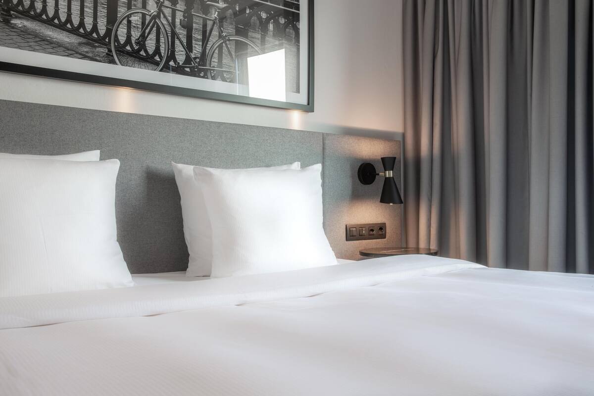 Habitación de hotel con una cama bien hecha, cabecera con panel gris claro, un cuadro colgado con bicicletas y una lámpara de pared.