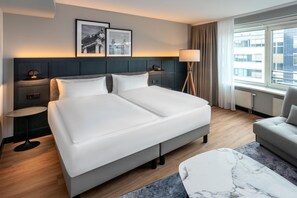 Un cuarto de hotel con una cama grande, una mesita de noche, una silla, una lámpara de pie y dos cuadros en la pared.