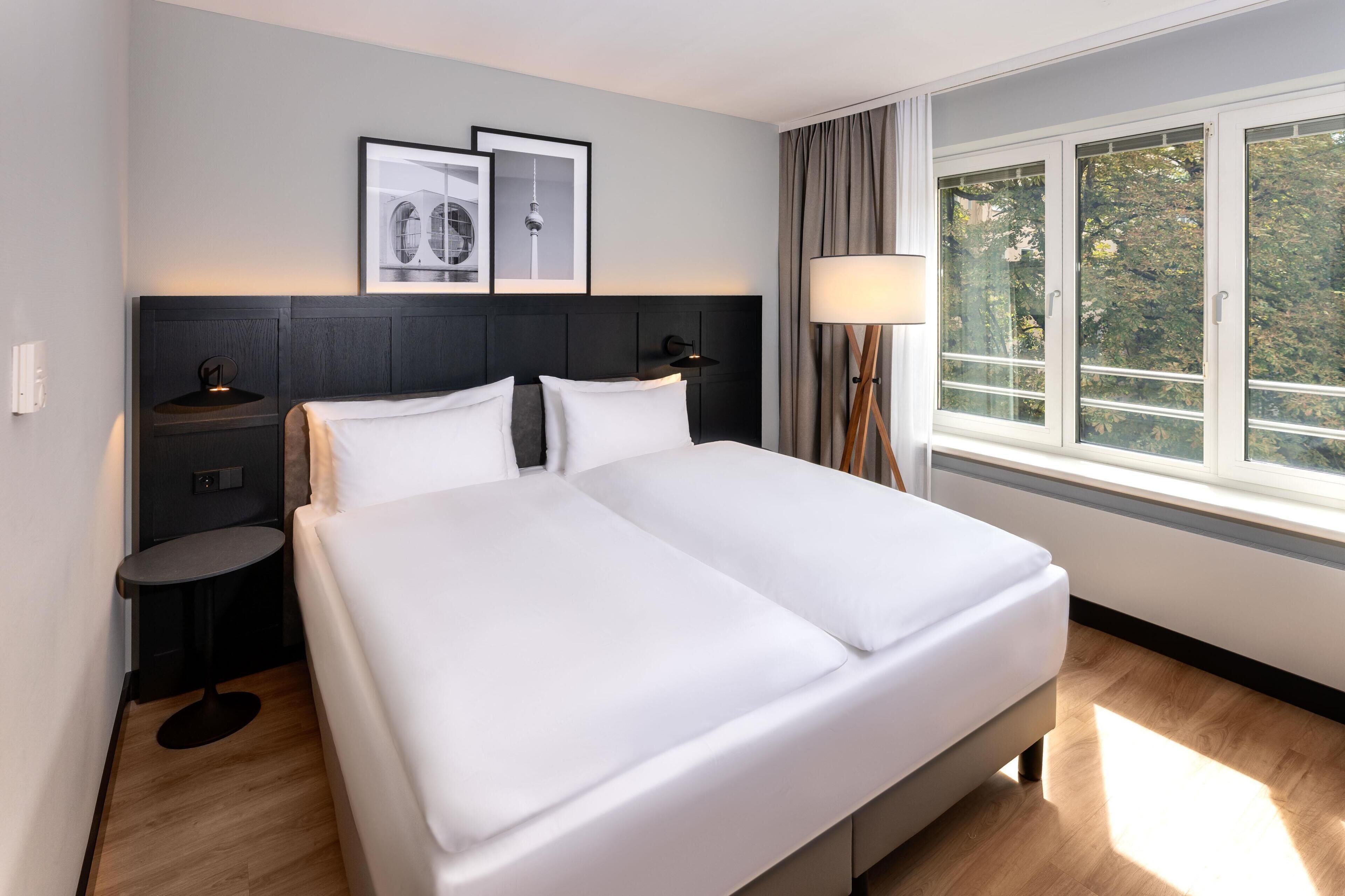 Ein Hotelzimmer mit Bett, Nachttischen, einer Lampe, einem Fenster mit Vorhängen und gerahmten Bildern an der Wand.