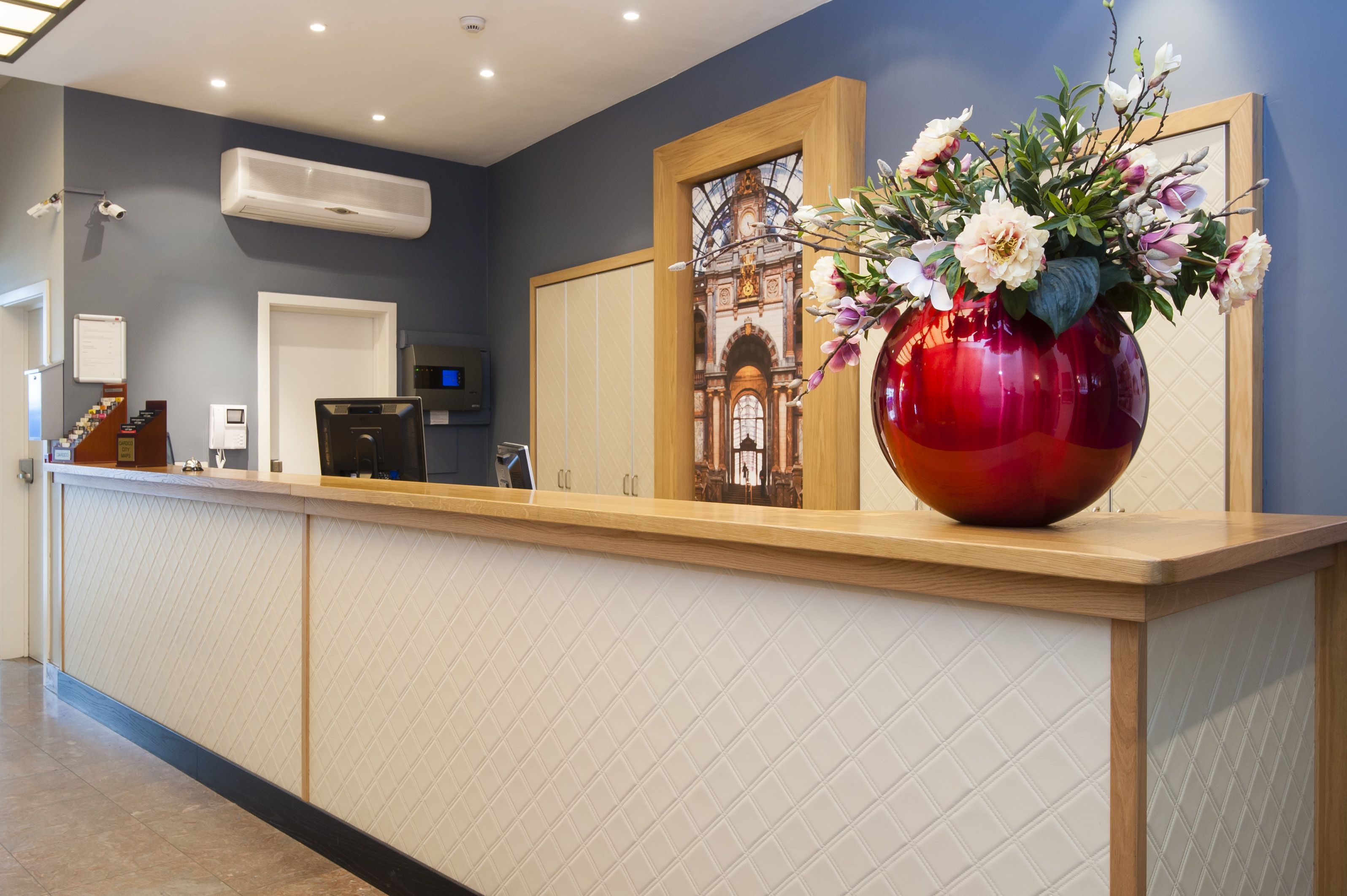 A reception desk with a large red vase of flowers, a digital screen, and a security camera.