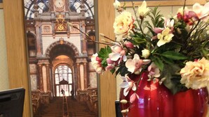 Una mesa de recepción con un gran jarrón rojo lleno de flores y un cuadro con la entrada de un edificio imponente.