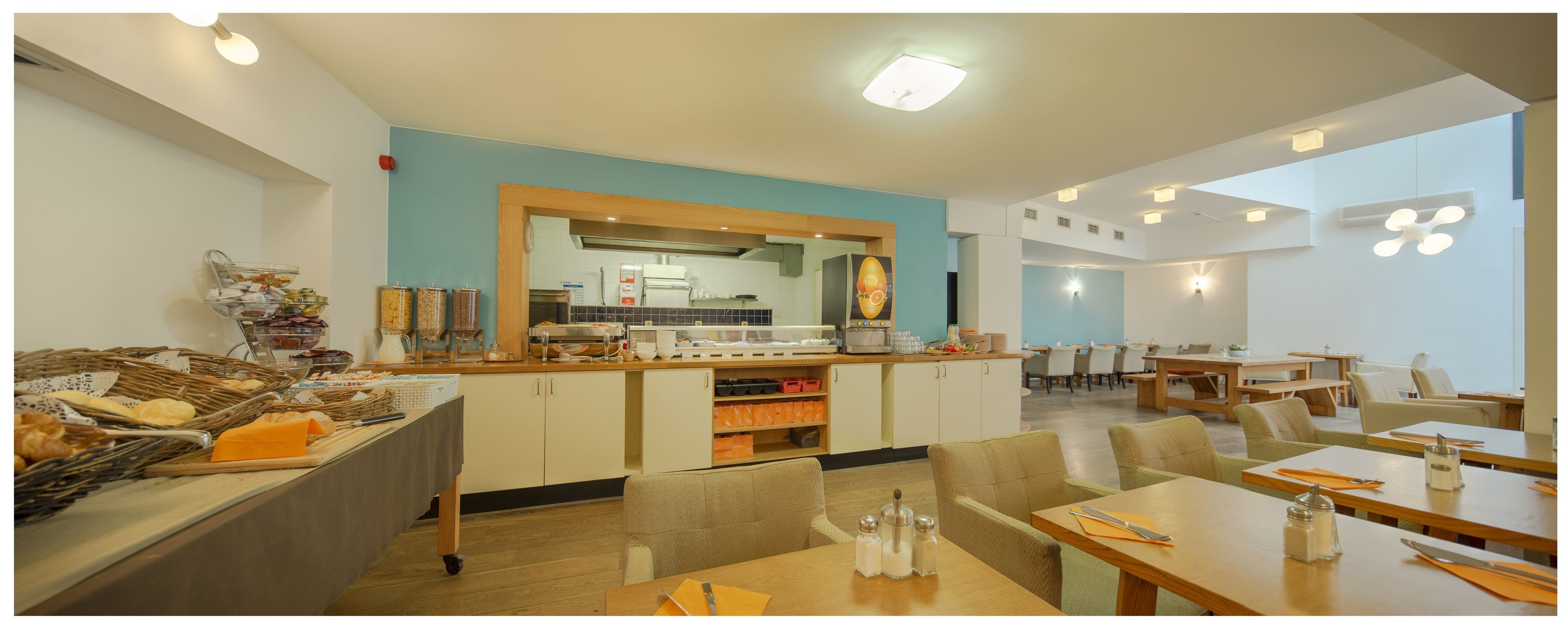 A modern dining area with wooden tables and chairs, a buffet with various food items, and a kitchen in the background.
