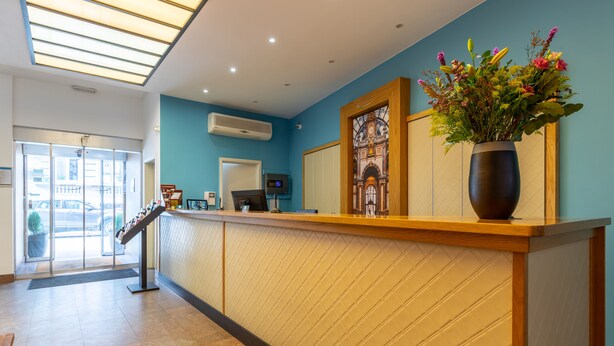 A reception area with a wooden counter, a vase of flowers, and a large window. - TripInn Eden Antwerp (Antwerp)