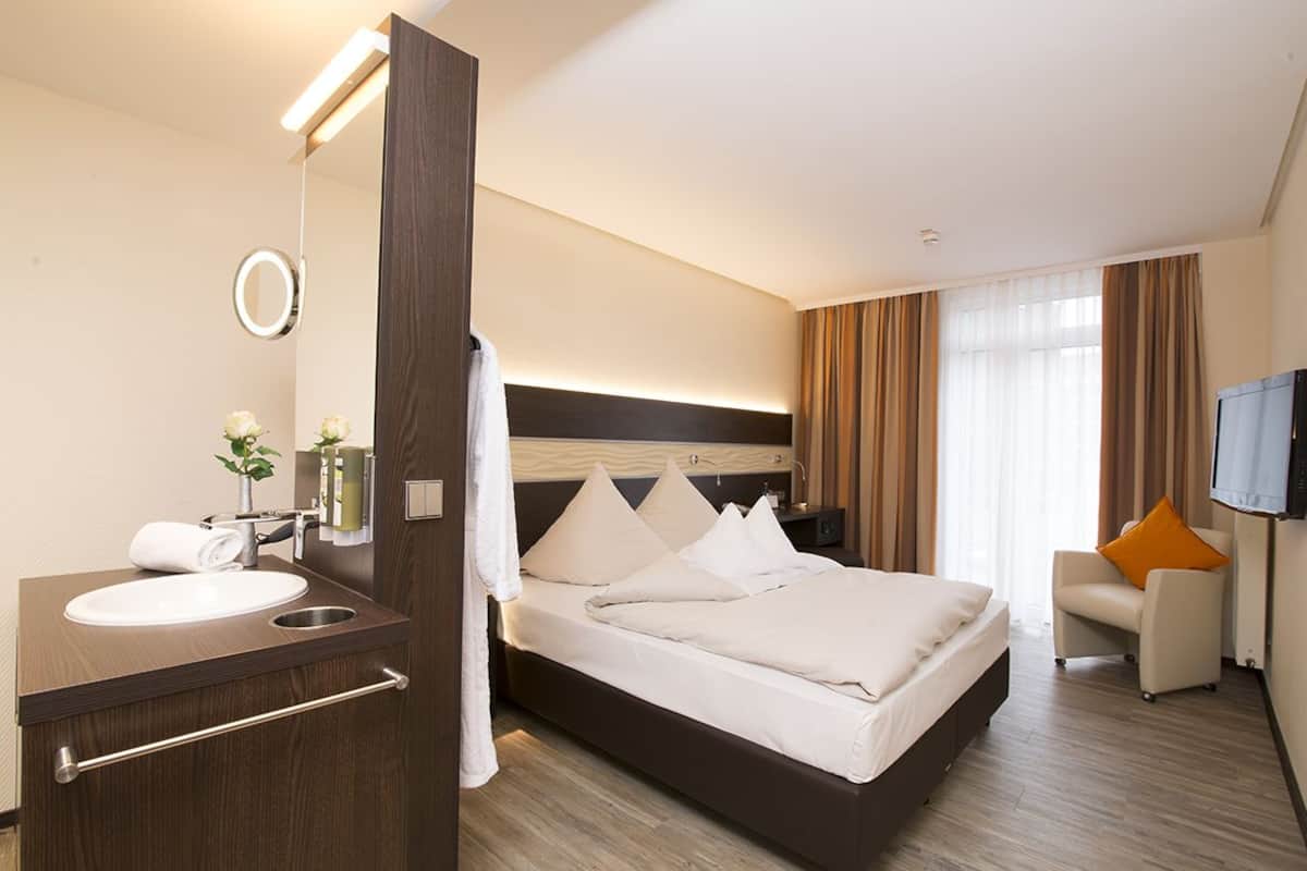 A hotel room with a large bed, a bedside table, a mirror, a TV, and a seating area with a chair and a sofa.