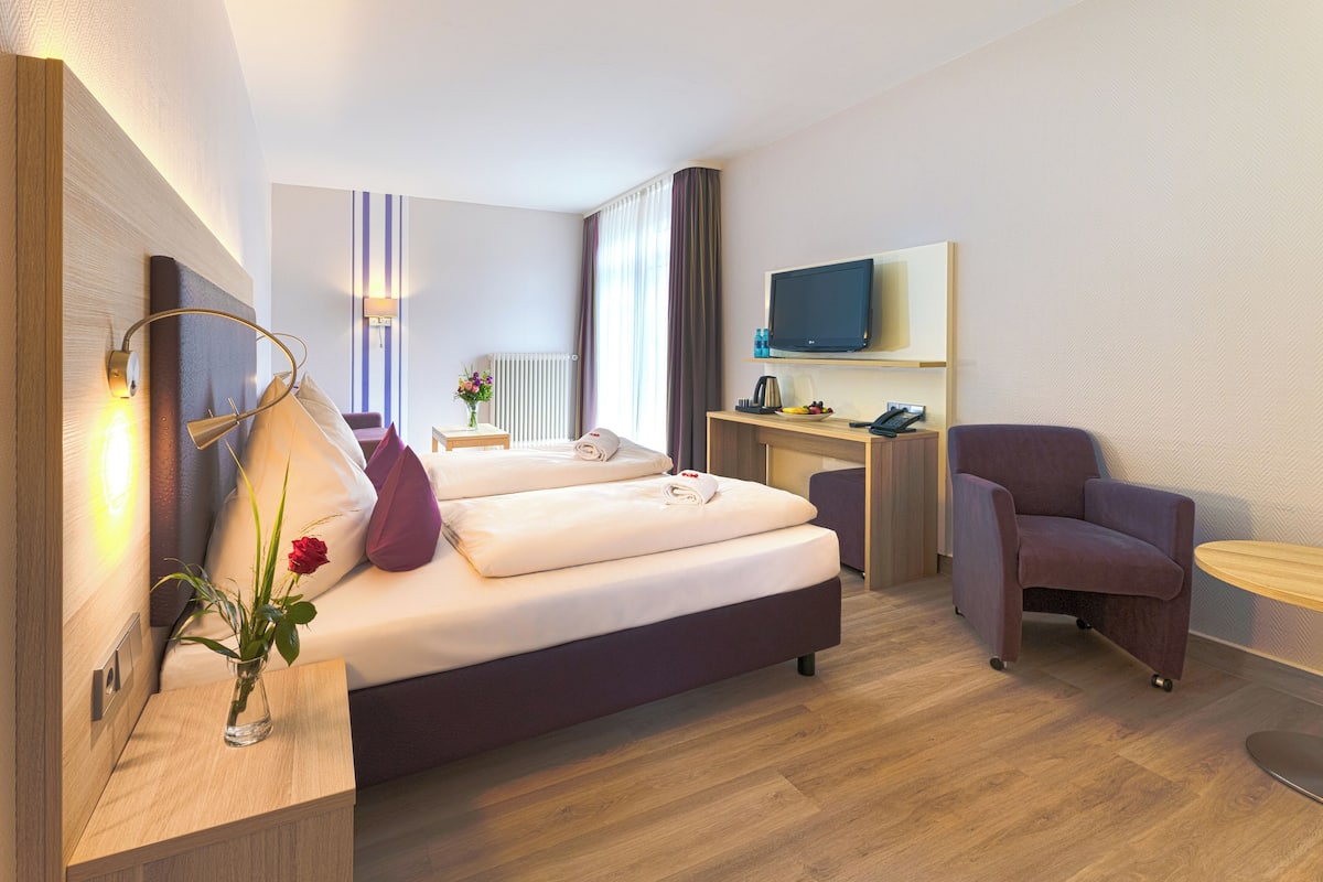 A modern hotel room with a bed, a desk, a chair, a TV, and a vase with flowers.