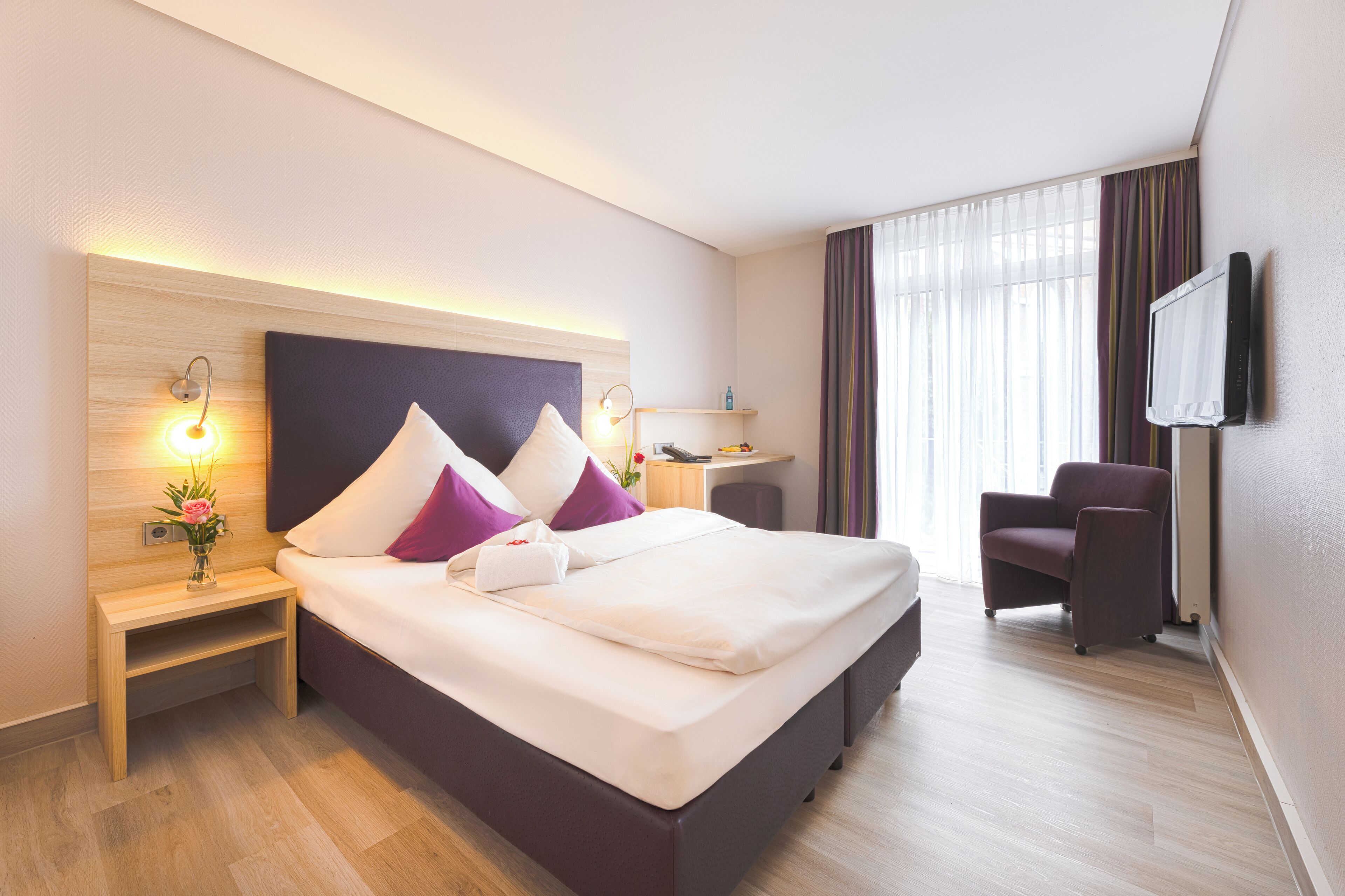 A hotel room with a large bed, bedside tables with lamps, a chair, a desk, and a television on the wall.