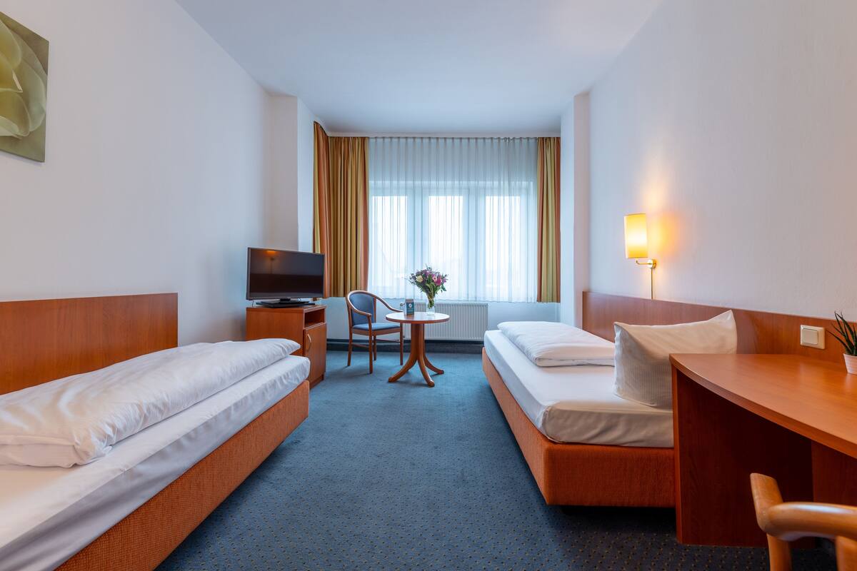 A hotel room with two single beds, a desk, a chair, a television, and a window with curtains.