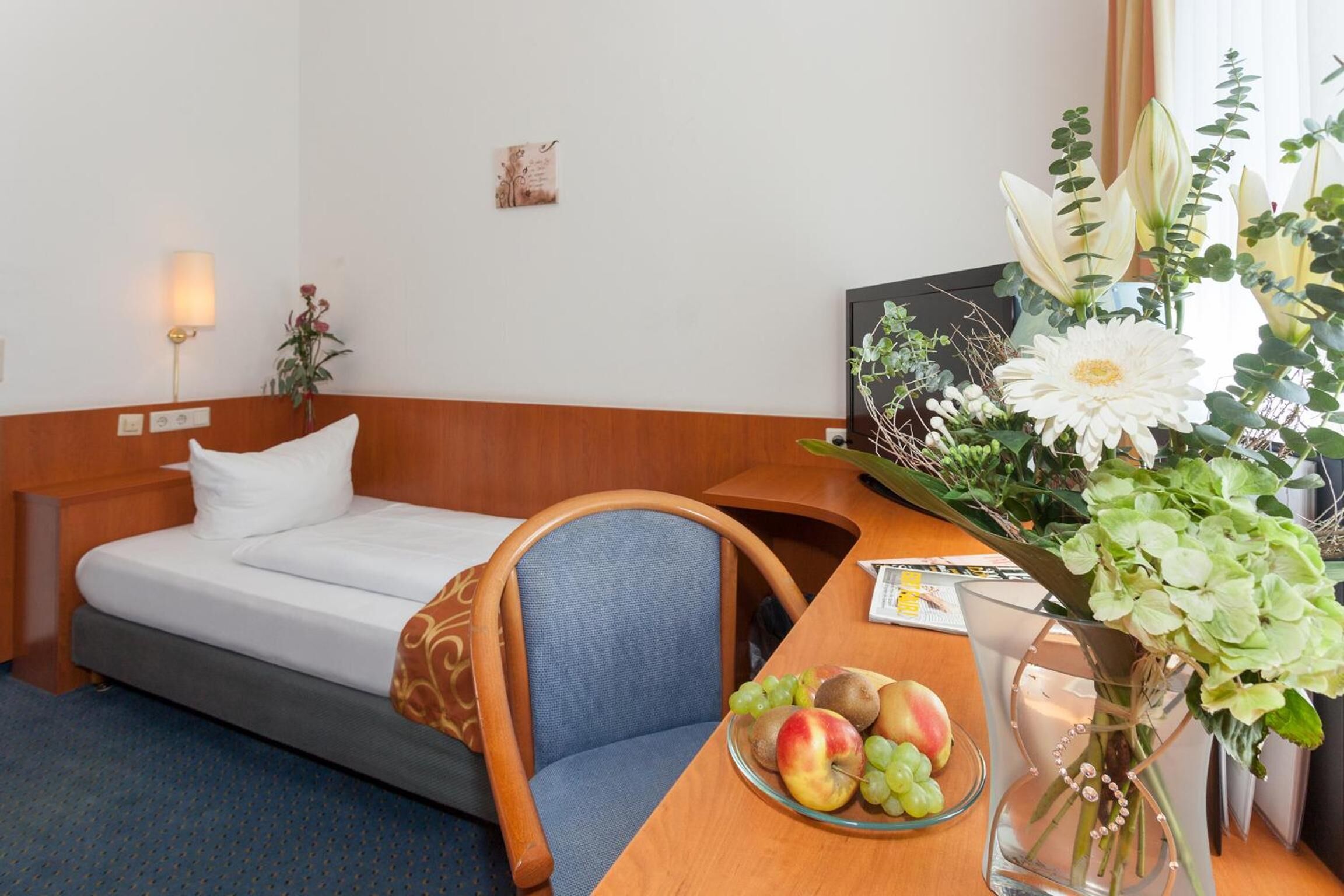 Ein Hotelzimmer mit einem Bett, einem hölzernen Schreibtisch mit Blumen und einer Obstschale.