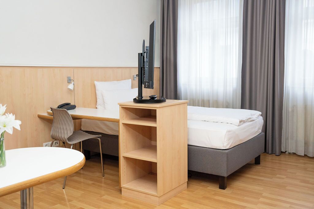 A hotel room with a bed, a desk with a chair, a TV on a stand, and a small table with a vase of flowers.