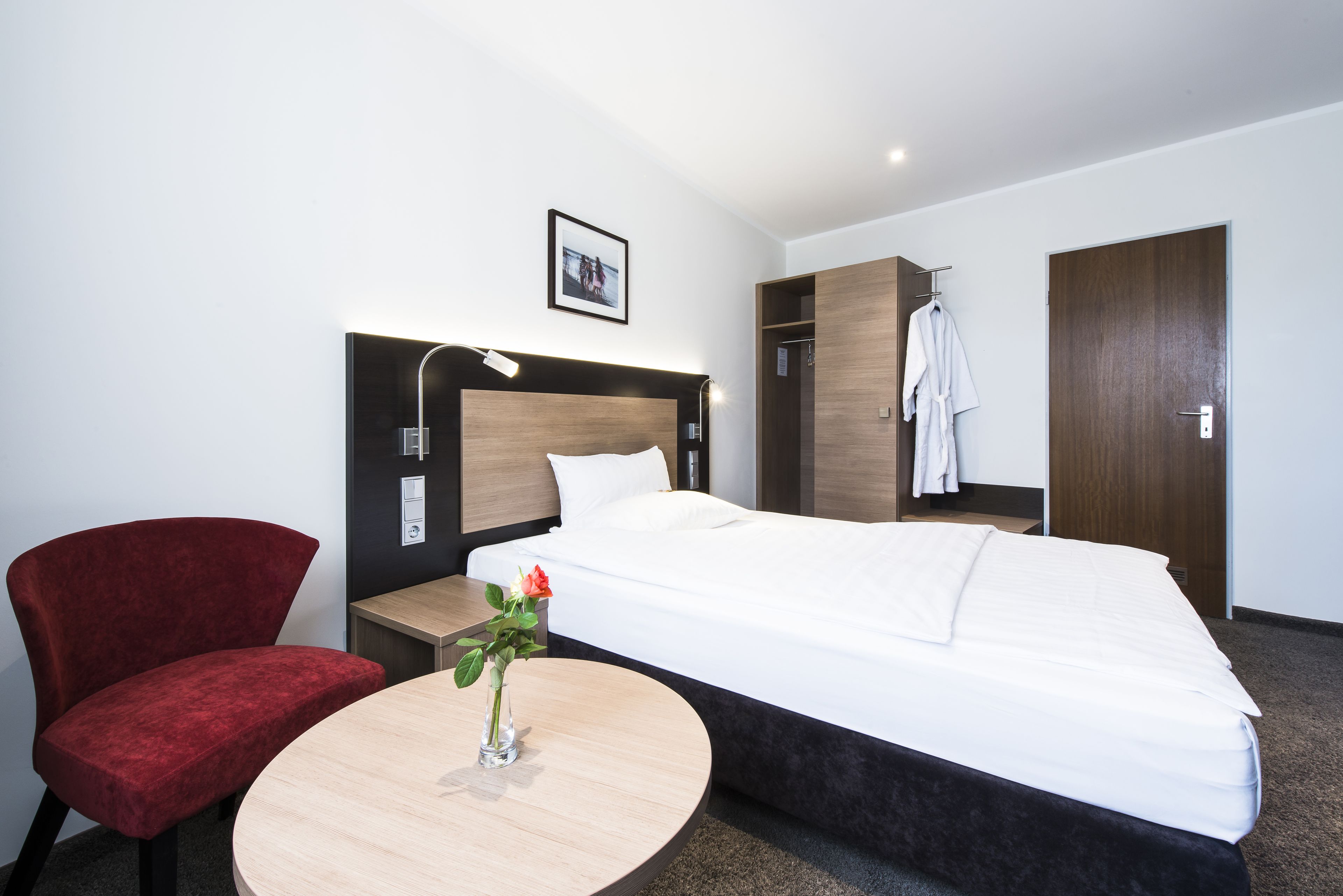 A hotel room with a bed, a red chair, a round table with a vase, and a wardrobe.